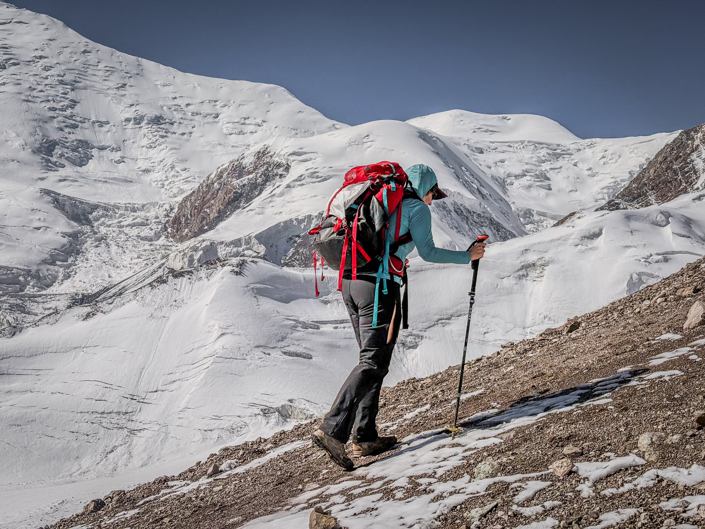 A climber with a red pack and trekking poles on a snowy moraine below the glaciers of Lenin Peak