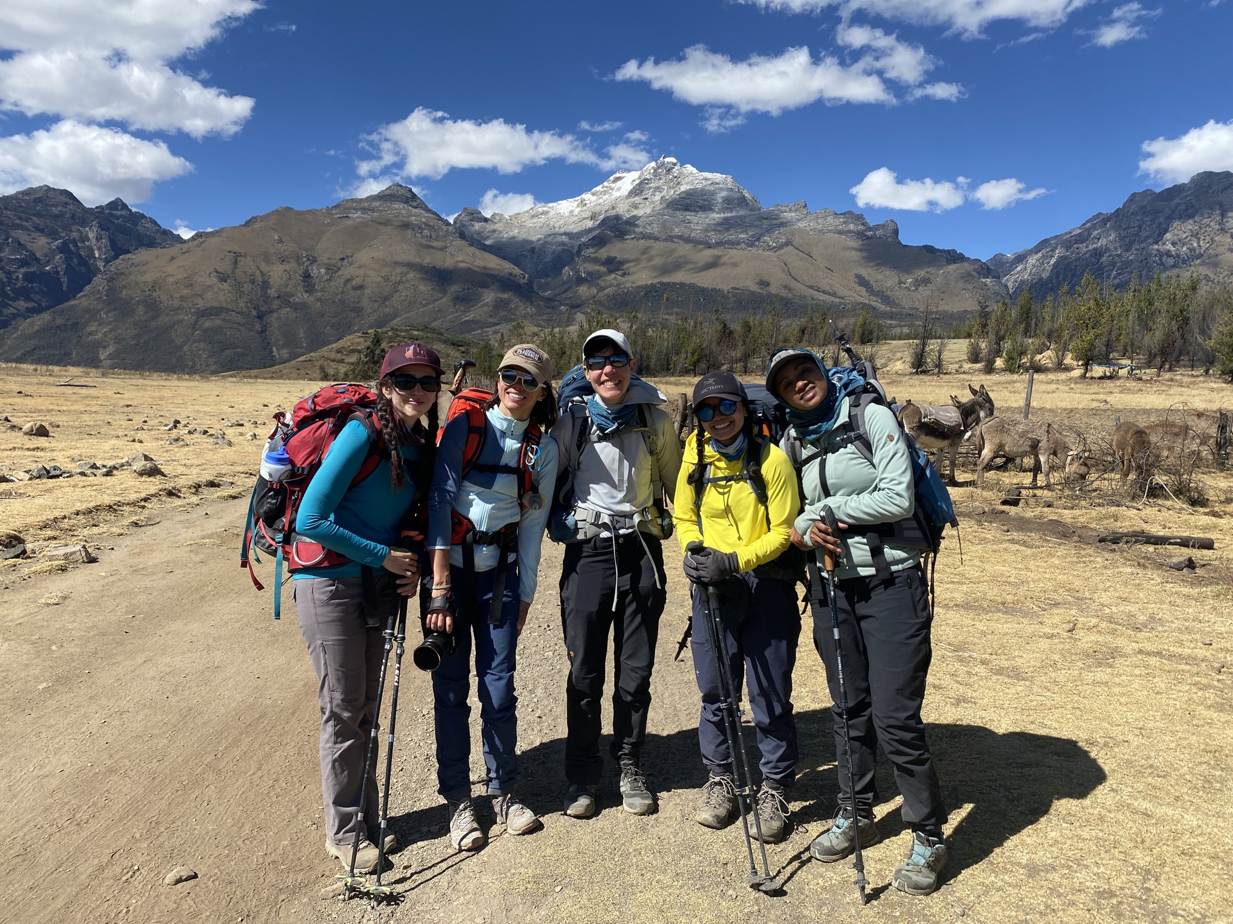 Five women with packs and trekking poles standing on a trail in Peru's Cordillera Blanca, a snowy peak behind them